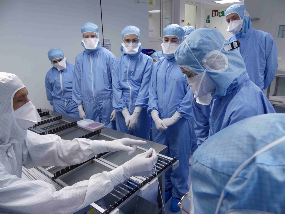 Girls' Day-Teilnehmerinnen und Mitarbeiter in Schutzkleidung beobachten eine Demonstration eines technischen Geräts in einem Reinraumlabor