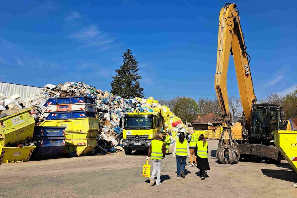 Vier Personen in gelben Warnwesten gehen auf eine große Müllhalde mit gelben Containern, einem Lastwagen und einem gelben Bagger zu.