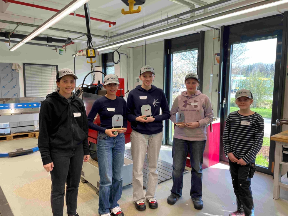 Fünf Girls' Day-Teilnehmerinnen mit grauen Caps stehen in einer Werkstatt, drei halten kleine Auszeichnungen in den Händen.