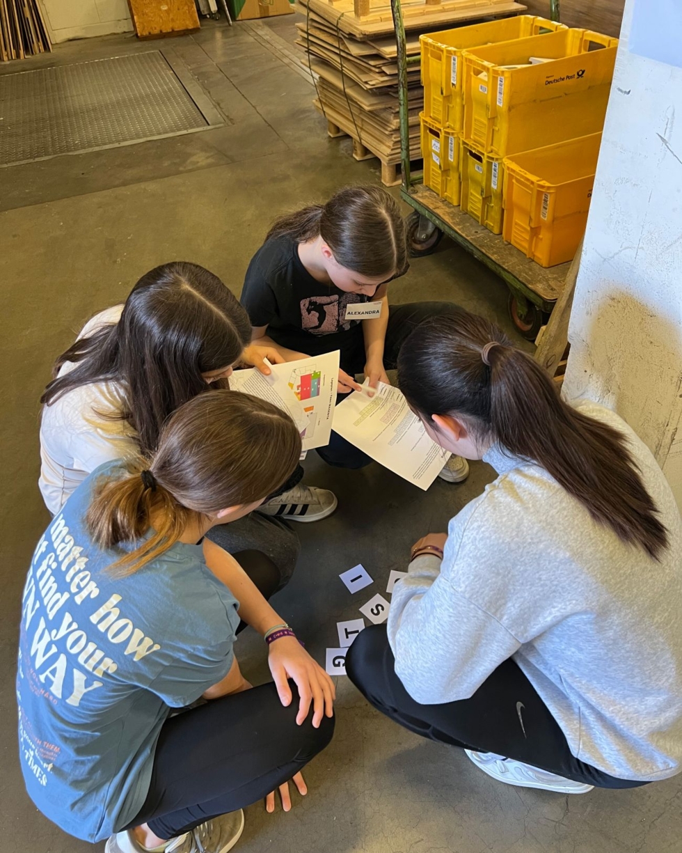 Vier Girls' Day-Teilnehmerinnen sitzen auf dem Boden in einer Lagerhalle und ordnen Buchstabenkarten, eine hält ein Heft mit farbiger Grafik