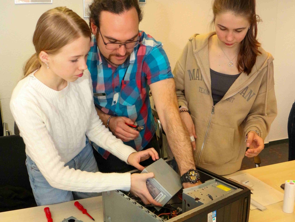 Drei Personen arbeiten gemeinsam an einem Computer. Eine junge Frau baut ein Bauteil aus, während ein Mann und eine weitere Frau unterstützen.