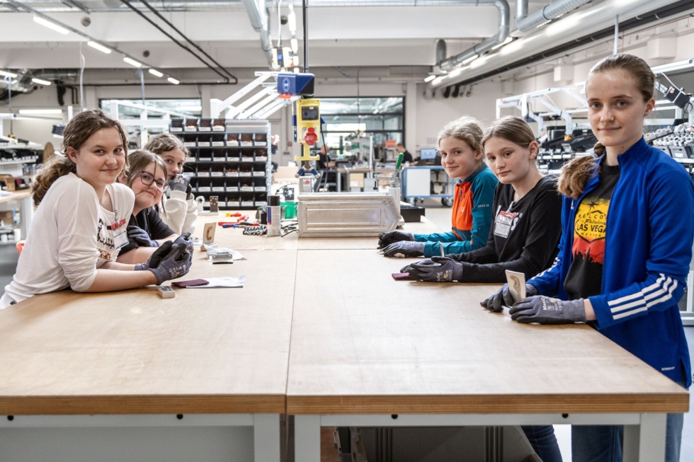 Sechs Girls' Day-Teilnehmerinnen arbeiten gemeinsam an einem Tisch in einer modernen Maschinenbauwerkstatt. Sie tragen Handschuhe