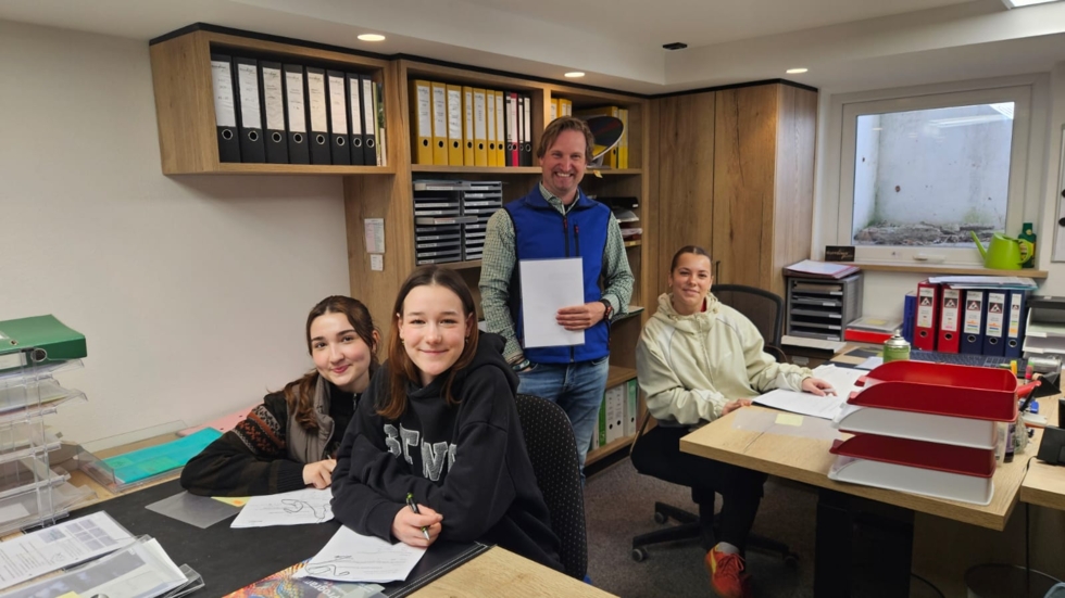 Drei Jugendliche und ein Mann in einem Büro. Die Jugendlichen sitzen an einem Tisch mit Unterlagen, der Mann steht hinter ihnen und hält ein Blatt Papier.