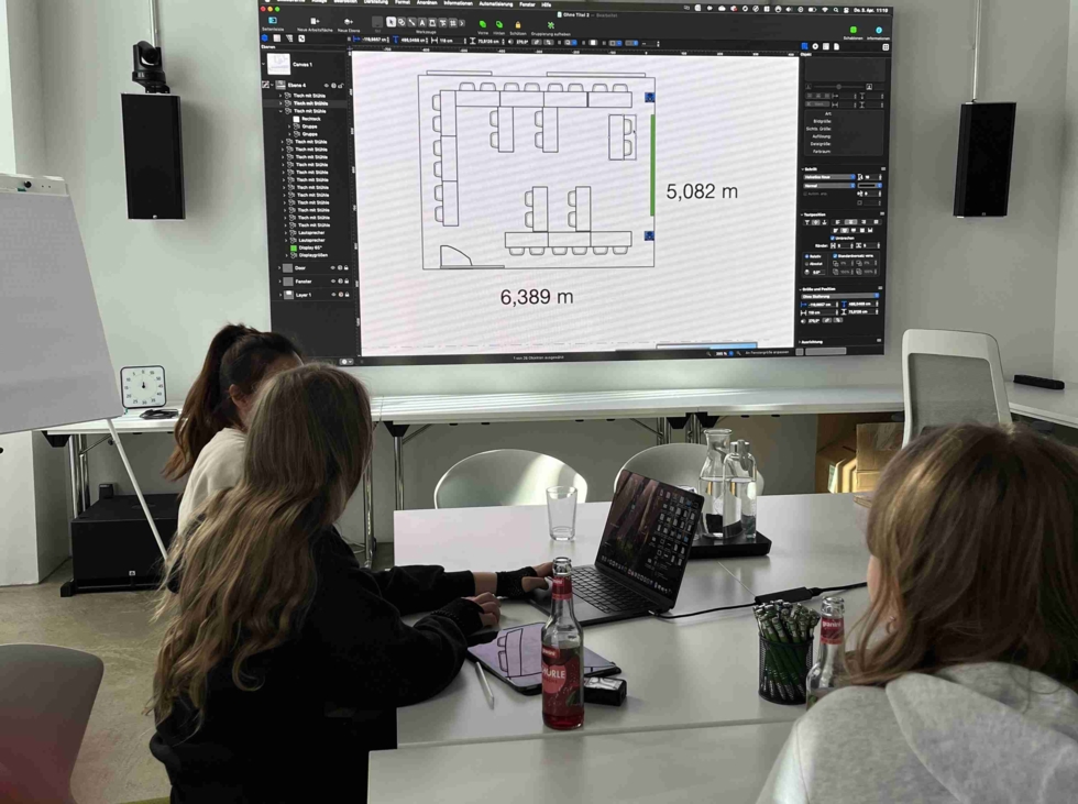 Drei Girls' Day Teilnehmerinnen sitzen an einem Tisch und betrachten einen großen Bildschirm mit einem AV-Plan