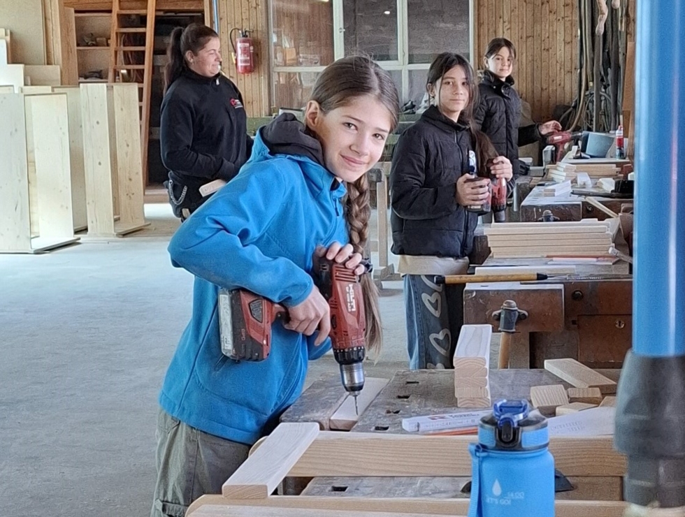 Ein Mädchen hält eine Bohrmaschine und lächelt. Im Hintergrund sind weitere Kinder und Werkbänke zu sehen.