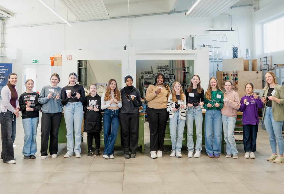 Gruppenfoto von zwölf Girls' Day-Teilnehmerinnen und zwei Mitarbeiterinnen in einer hellen Werkstatt, alle lächeln und halten kleine Herzformen aus Draht in den Händen