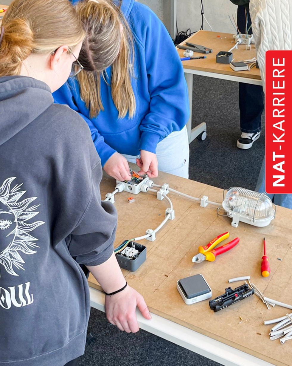 Zwei Girls' Day-Teilnehmerinnen arbeiten an einem Tisch an einer elektrischen Schaltung mit Kabeln, Steckdosen und Werkzeugen wie Zange und Schraubenzieher