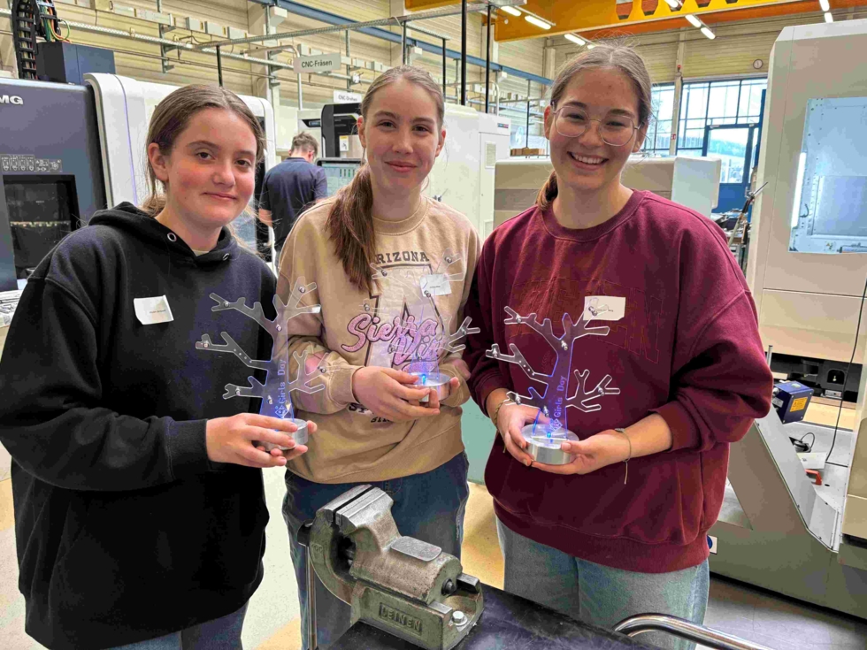 Drei Girls' Day-Teilnehmerinnen in Werkstatt halten beleuchtete Metallbäume als Auszeichnungen, im Hintergrund Maschinen und Werkstattumgebung
