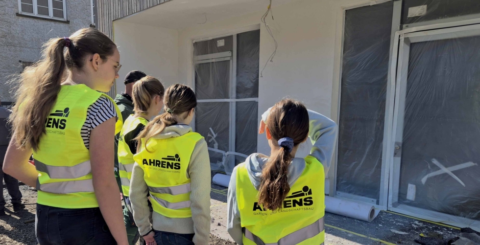 Girls' Day-Teilnehmerinnen stehen mit gelben Warnwesten vor einem Gebäude mit abgedeckten Fenstern und Türen, vermutlich bei einer Baustellenbesichtigung