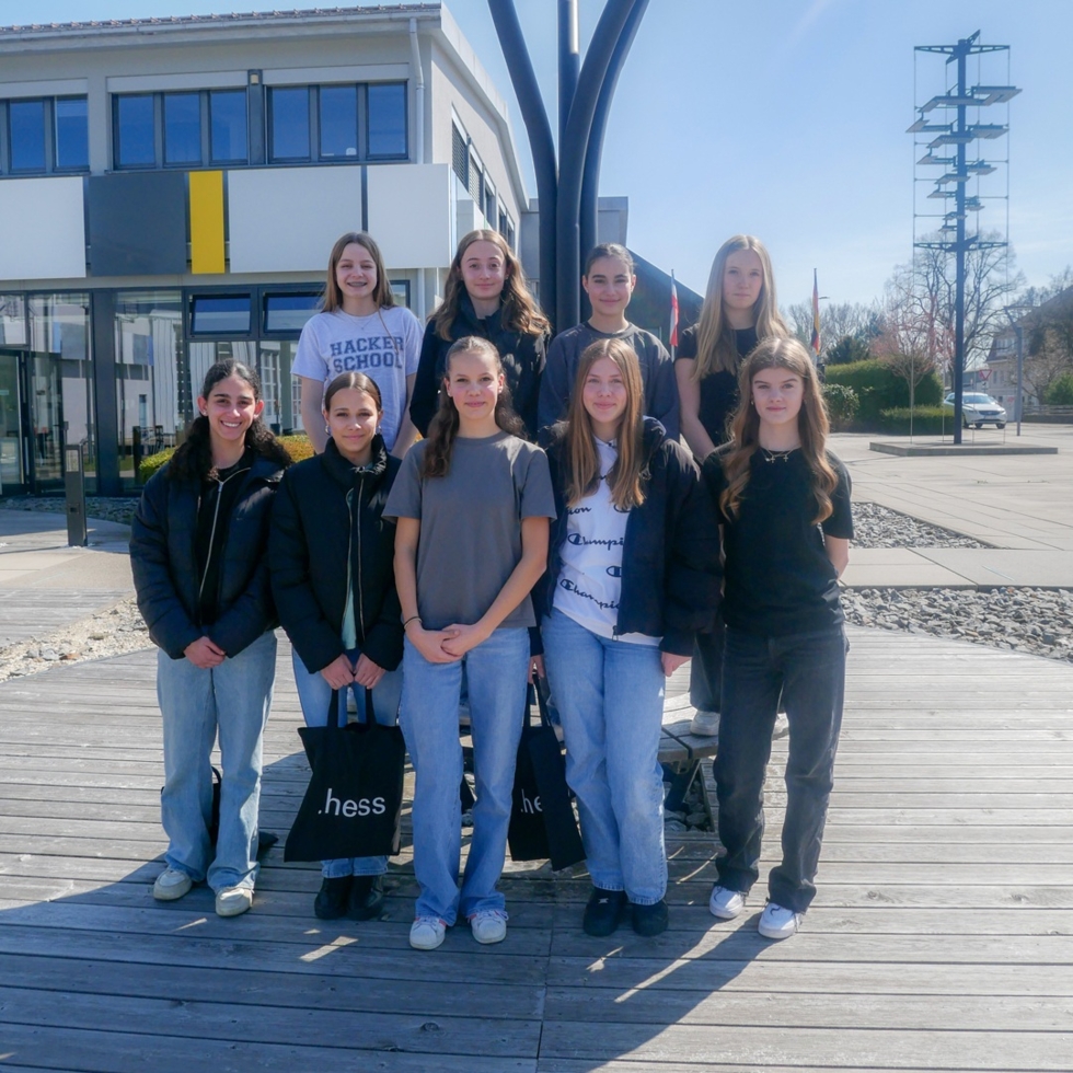 Neun Girls' Day-Teilnehmerinnen stehen in zwei Reihen auf einem Holzsteg vor einem modernen Gebäude mit gelbem Akzent