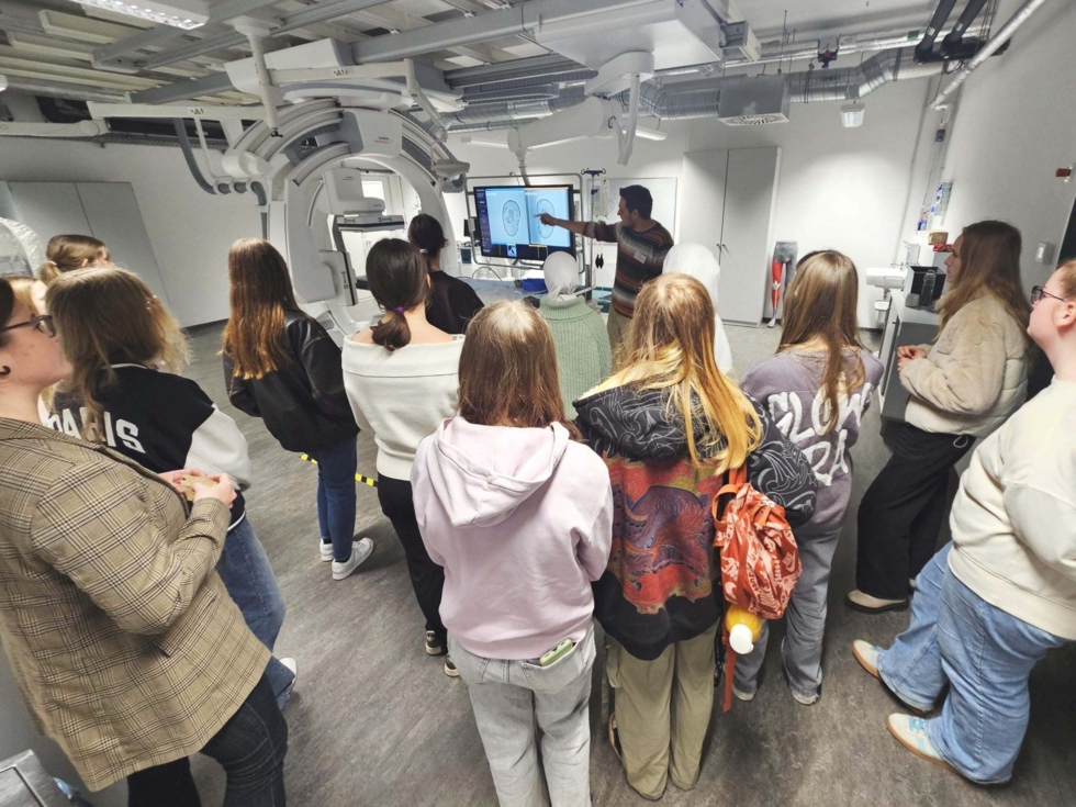 Gruppe von Girls' Day-Teilnehmerinnen in einem Labor, die einem Mitarbeiter zuhören, der auf einen Bildschirm mit technischen Zeichnungen zeigt