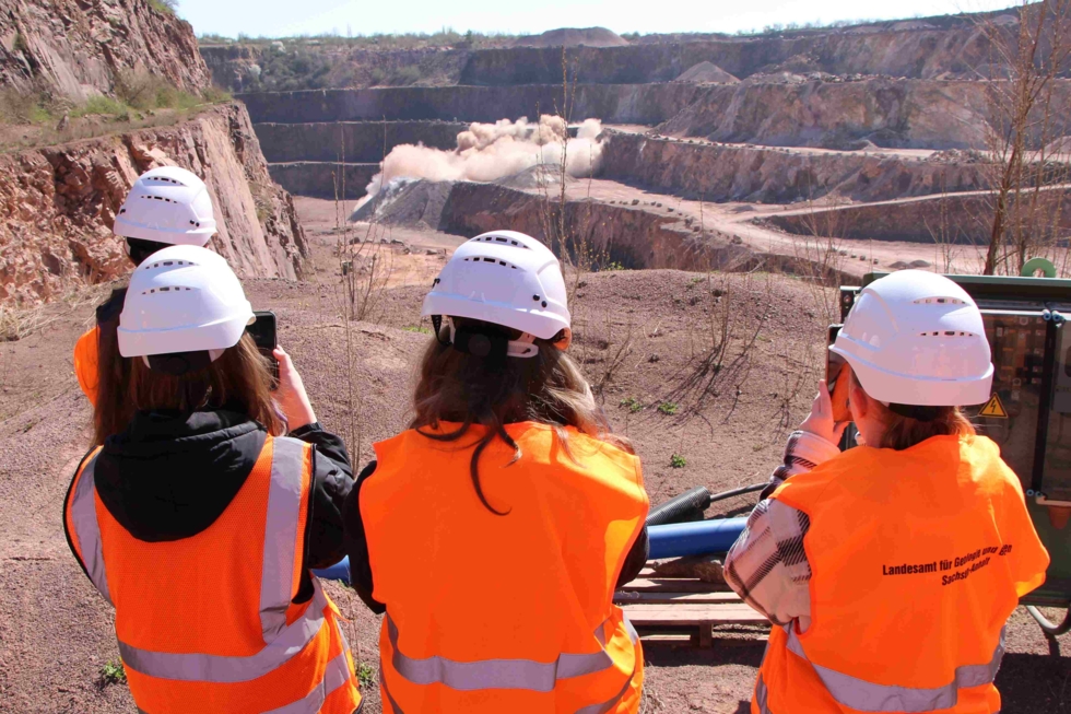 Gruppe von vier Personen in orangefarbenen Sicherheitswesten und Helmen, die eine Sprengung in einem Steinbruch beobachten.