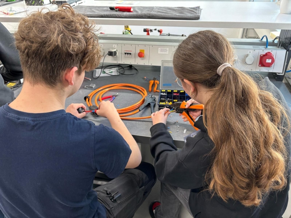 Mitarbeiter und Girls' Day-Teilnehmerin arbeiten an einem Tisch mit Werkzeugen und orangefarbenen Kabeln in einer Werkstatt