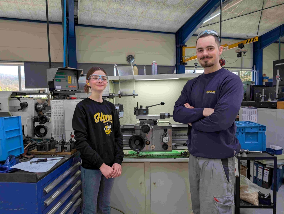 Girls' Day-Teilnehmerin und ein Ausbilder stehen in einer Werkstatt; im Hintergrund sind Maschinen und Werkzeuge sichtbar