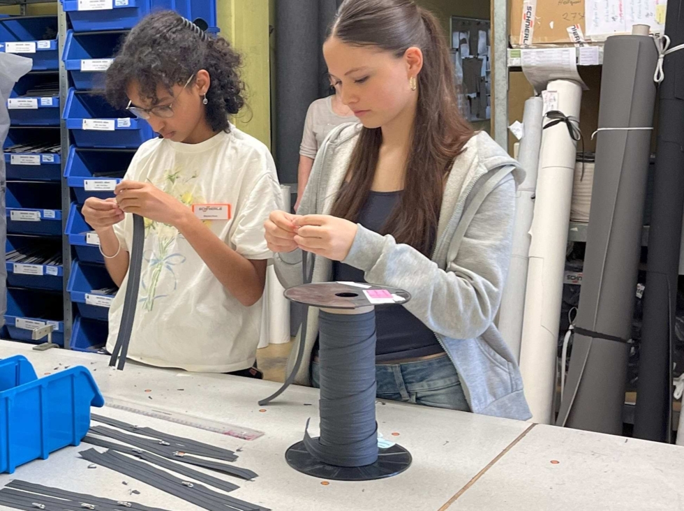 Zwei Girls' Day-Teilnehmerinnen arbeiten an einem Tisch mit schwarzen Kabelbindern in einem Lagerraum mit Regalen und blauen Aufbewahrungsboxen