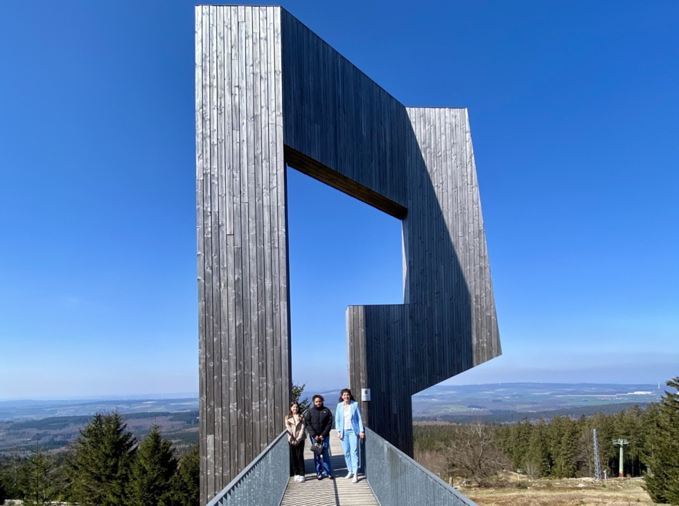 Drei Personen stehen auf einer Aussichtsplattform vor einer großen, modernen Holzskulptur in Form eines Rahmens, umgeben von einer Landschaft unter blauem Himmel.