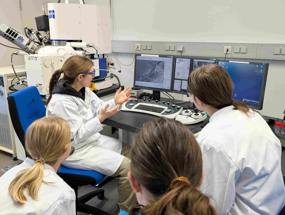 Technikerin erklärt den Teilnehmerinnen des Girls' Day die Funktionsweise eines Rasterelektronenmikroskops, während sie auf die Bildschirme schauen.