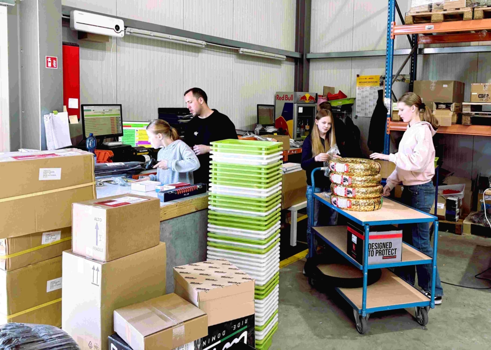 Drei Girls' Day-Teilnehmerinnen und ein Mitarbeiter verpacken und sortieren Waren in einem Lager mit Kartons, Regalen und einem Wagen mit verpackten Produkten