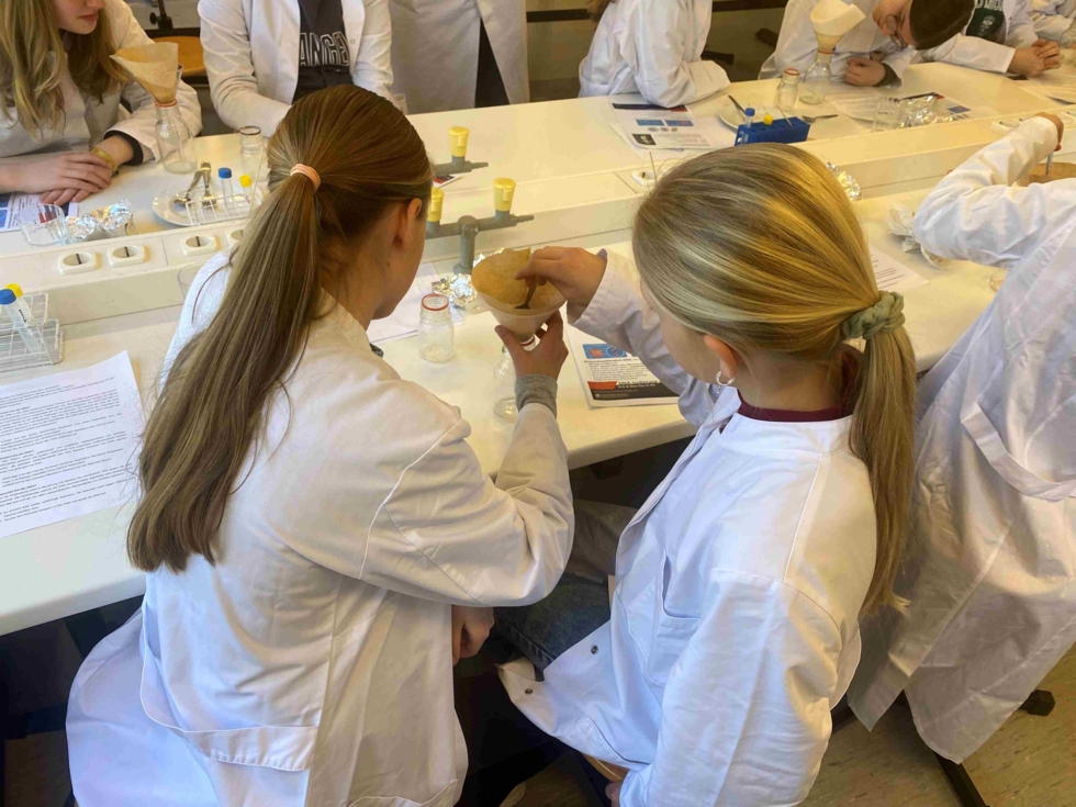 Zwei Girls' Day-Teilnehmerinnen in weißen Laborkitteln arbeiten gemeinsam an einem Experiment an einem Labortisch mit verschiedenen Geräten und Materialien